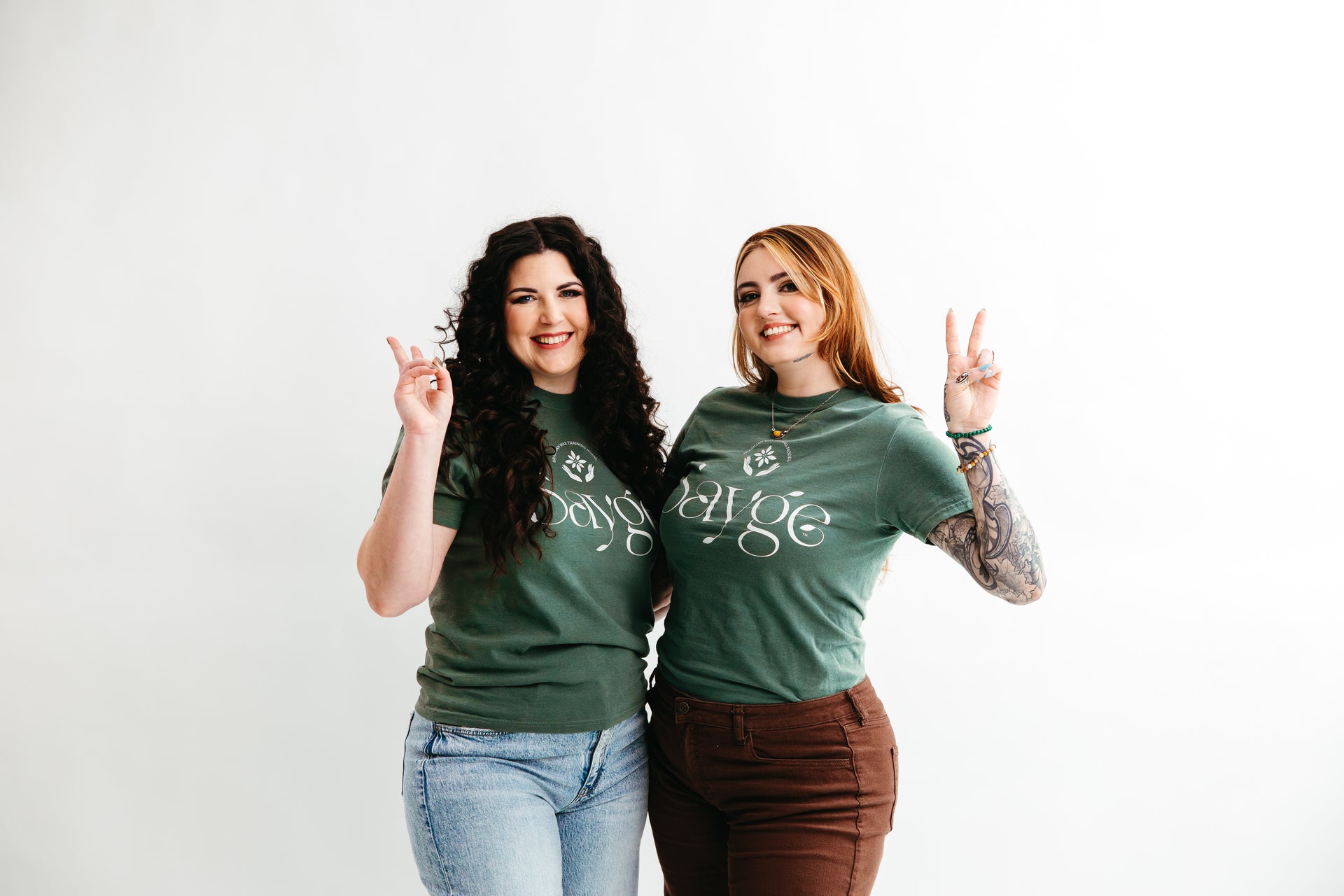 Founders Cadie and Alyssa wearing green t-shirts with a white Sayge logo, making peace signs against a white background